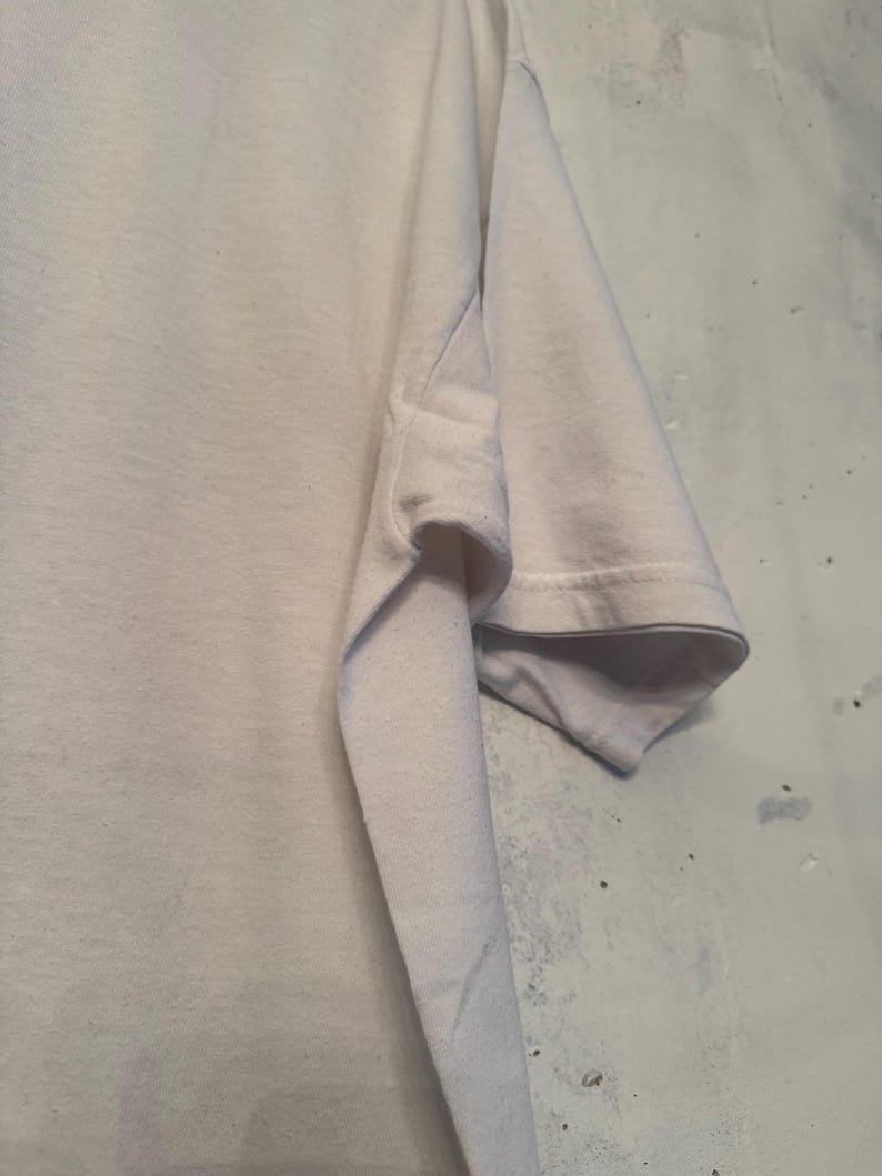以下が含まれることがあります： 白いTシャツの袖のクローズアップ。 袖口で折りたたまれ、内側の生地が見えています。 生地は柔らかく、軽量な素材のようです。 背景は、質感のある明るい色の壁です。