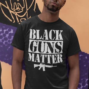 Può includere: Una t-shirt nera a maniche corte con la scritta bianca in stile vintage "BLACK GUNS MATTER" stampata in grassetto sul petto, accompagnata da un'illustrazione grafica bianca di un fucile posizionata direttamente sotto il testo.