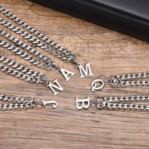 Puede incluir: Collares de cadena plateados con colgantes individuales de letras que deletrean las letras J, N, A, M, Q y B. Los collares se muestran sobre una superficie de madera, con un estuche negro en el fondo.