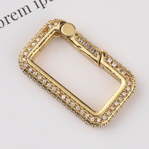 May include: A gold-toned metal clasp with a square shape and a decorative border of small clear rhinestones.