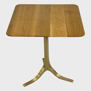 Puede incluir: Una tapa de mesa cuadrada de madera con un acabado de madera natural se asienta sobre una base de metal dorado con tres patas. La base tiene un diseño curvo.