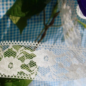 May include: A strip of white lace trim featuring a delicate floral pattern with a scalloped edge and a fine hexagonal mesh background. The lace is woven with a consistent thread density, showcasing intricate petal details and a structured top border.
