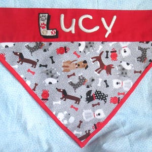 May include: Red and gray dog bandana with the name "Lucy" embroidered in white. The bandana features a pattern of various dog breeds, bones, and fire hydrants. The top section is solid red.