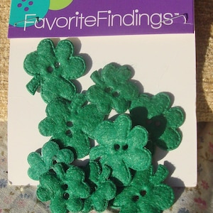 Puede incluir: Un paquete de botones FavoriteFindings con forma de tréboles verdes. Cada botón tiene dos agujeros en el centro para su fijación. Los botones están sujetos a una tarjeta blanca con un encabezado morado que muestra el nombre de la marca en blanco.