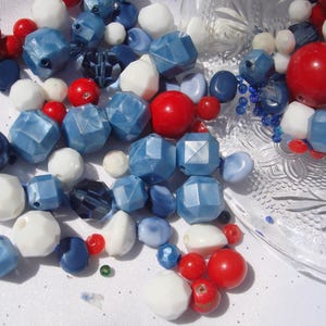 May include: A collection of red, white, and blue beads in various shapes and sizes. The beads include round, faceted, and irregular shapes, with a mix of glossy and matte finishes. Some beads are scattered on a white surface, while others are in a clear glass dish.