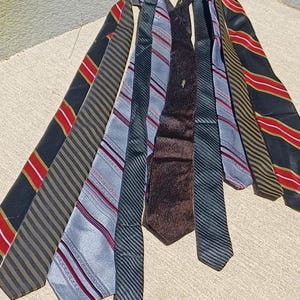 Puede incluir: Una colección de siete corbatas dispuestas en forma de abanico. Las corbatas presentan varios patrones, incluyendo rayas diagonales, tela texturizada de color marrón oscuro sólido y rayas finas en colores negro, rojo, plata, gris y oro.