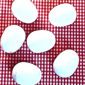 May include: Five smooth, rounded, white stones arranged in a scattered pattern. Each stone has an opaque, matte white surface with a solid, uniform appearance. The stones are oval and circular in shape, varying slightly in size and orientation.