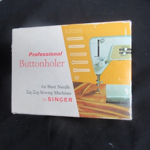 Puede incluir: Una caja vintage para una ojaladora profesional Singer para máquinas de coser zig-zag de aguja inclinada. La caja tiene un diseño blanco y naranja con una imagen de una máquina de coser. El texto "Professional Buttonholer" es rojo.