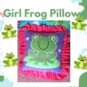 May include: A square purple pillow featuring a green frog appliqué sitting on a light green lily pad. The pillow is bordered by a ruffled hot pink fabric trim. The name Amberly is embroidered in purple thread on the frog's belly. The text Girl Frog Pillow appears above.