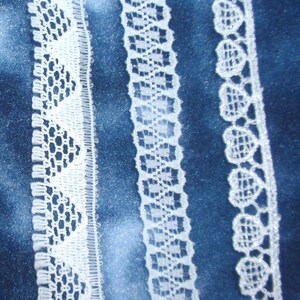 May include: Three strips of white lace trim arranged vertically. The left strip features a repeating triangular pattern, the middle strip has a geometric grid design, and the right strip displays a series of small, connected heart-shaped motifs.