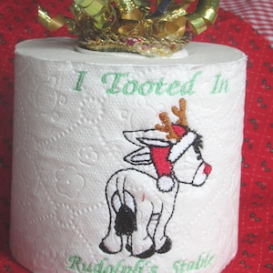 Könnte beinhalten: Eine weiße Toilettenpapierrolle mit einer Stickerei eines Esels, der eine Weihnachtsmütze und ein Rentiergeweih trägt. Der Text "I Tooted In" steht über dem Esel und "Rudolph's Stable" ist darunter in grünem Garn eingestickt.