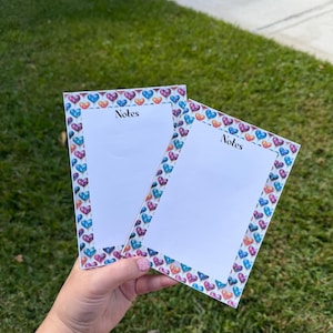 May include: Two rectangular notepads with a white interior and a border of colourful hearts. The word "Notes" is printed in black at the top of each notepad. The notepads are held against a background of green grass.