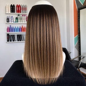 May include: A long, straight hair extension piece with a dark brown to light brown ombre effect. The hair is styled with a smooth, sleek finish, and the color transition is gradual. The hairpiece is designed to be worn on the head.