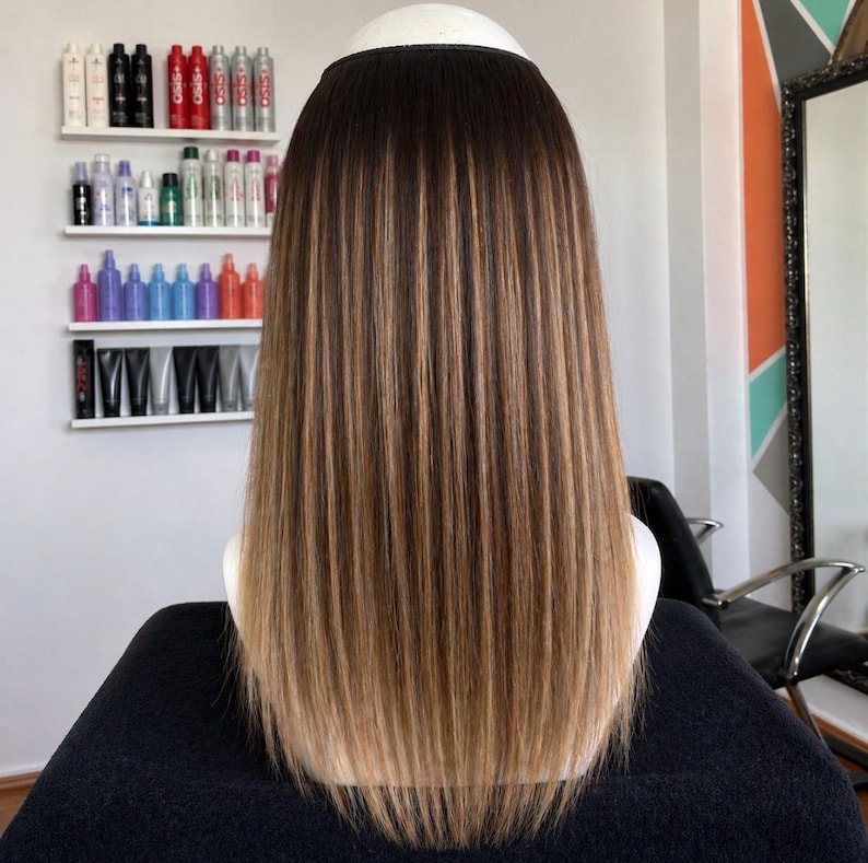 May include: A long, straight hair extension piece with a dark brown to light brown ombre effect. The hair is styled with a smooth, sleek finish, and the color transition is gradual. The hairpiece is designed to be worn on the head.