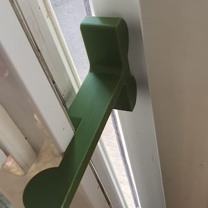 Op de afbeelding: Een groene, 3D-geprinte raamvergrendeling geïnstalleerd op een wit raamkozijn. De vergrendeling heeft een solide, hoekige vorm met een gebogen uiteinde, ontworpen om een raam te beveiligen. Het raam heeft een scherm en is gedeeltelijk open.