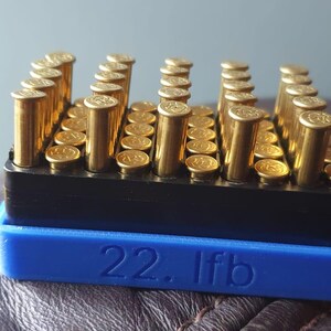 Peut inclure: Un support de munitions noir et bleu rempli de cartouches de calibre .22 long rifle de couleur laiton. La base bleue porte l'inscription "22. lfb". Les cartouches sont disposées en rangées.