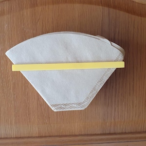 Può includere: Una pila di filtri per caffè in carta beige tenuti insieme da un supporto in plastica giallo. I filtri sono triangolari e presentano un motivo strutturato sui bordi. Il supporto è una sottile barra rettangolare.