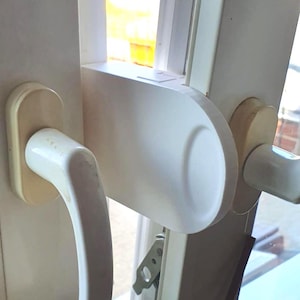 Op de afbeelding: Een witte kunststof raambeveiliging gemonteerd tussen twee witte raamkozijnen. Het apparaat is voorzien van een gebogen, ergonomische handgreep en een plat, afgerond vergrendelingsmechanisme om het raam vast te zetten.