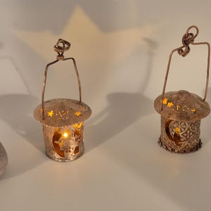 May include: Two decorative metal lanterns with intricate cut-out designs. Each lantern has a copper-colored handle and a warm, glowing light inside. The lanterns are cylindrical with a domed top, creating a cozy ambiance.