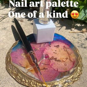 May include: A nail art palette with the text "Nail art palette One of a kind". The palette is a mix of pink, gold, and blue, with a gold rim. A nail polish bottle and a nail art brush are on the palette.