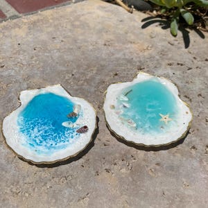 May include: Two seashell-shaped resin art pieces. Each features a vibrant blue and white ocean scene with small shells and a gold-colored rim. One has a starfish. The pieces are on a textured surface.