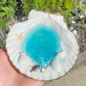 May include: A decorative seashell dish with a gold-painted rim. The interior features a turquoise resin pool with sand, a starfish, and small seashells. A silver turtle charm adds detail to the beach scene.