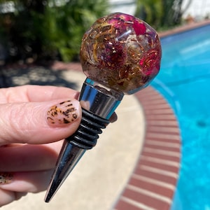 May include: A decorative wine bottle stopper. The clear, spherical top is filled with dried flowers and gold flakes. It has a silver metal base and a black rubber seal. The background shows a swimming pool.