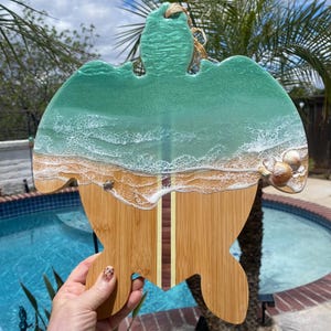 Può includere: Tagliere a forma di tartaruga con un design a scena di spiaggia. La parte superiore presenta un oceano in resina turchese con onde bianche e schiumose. Il fondo è in bambù con una striscia di tavola da surf. Conchiglie sono incorporate sul lato destro.