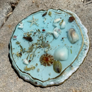 Puede incluir: Un posavasos redondo de resina azul claro con conchas marinas, una estrella de mar y copos de oro incrustados. El posavasos tiene un borde dorado y un borde blanco texturizado. Un artículo decorativo para la decoración costera.