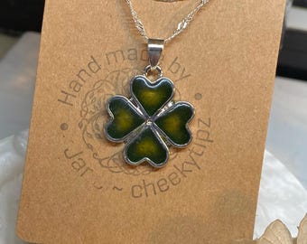 May include: Silver-toned necklace featuring a four-leaf clover pendant with green enamel filling. The pendant is on a delicate chain, displayed on a brown card with the text "Hand made by Jar ~ cheekytipz". The background includes dried flowers.