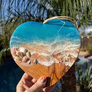 Puede incluir: Una placa de madera en forma de corazón con un diseño de resina que representa una escena de playa con agua turquesa, olas de espuma blanca y arena. La superficie está decorada con varias conchas marinas pequeñas y una estrella de mar.
