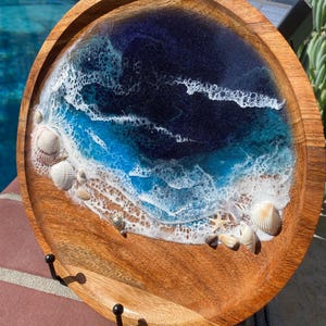 Può includere: Un vassoio rotondo in legno con un design oceanico in resina. Il vassoio presenta una scena oceanica blu scuro e turchese con onde bianche e schiumose e conchiglie. Tre ganci neri sono attaccati al bordo del vassoio.