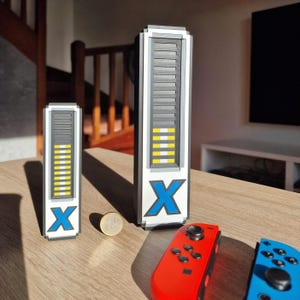 May include: Two white, gray, and yellow pixelated battery-shaped objects with a blue "X" logo, next to a coin and red and blue game controllers. The objects have a segmented design, with a series of horizontal lines and yellow bars indicating battery level.