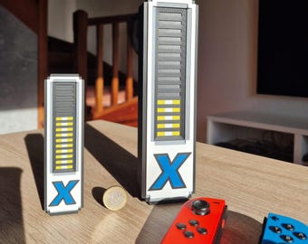 May include: Two white, grey, and yellow pixelated battery-shaped objects with a blue "X" logo, next to a coin and red and blue game controllers. The objects have a segmented design, with horizontal lines and yellow bars indicating battery level.