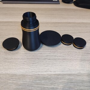 May include: A black bottle with a cylindrical neck and a rounded body, accented with two thin orange stripes. Several black, circular lids of varying sizes are arranged around the bottle, suggesting a set of containers or closures.
