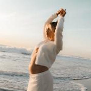 K&ouml;nnte beinhalten: Eine Frau in einem wei&szlig;en, kurz geschnittenen Oberteil mit langen &Auml;rmeln und einem passenden Rock steht an einem Strand. Die Meereswellen sind im Hintergrund unter einem hellblauen Himmel zu sehen. Die Frau hat die Arme &uuml;ber dem Kopf erhoben.