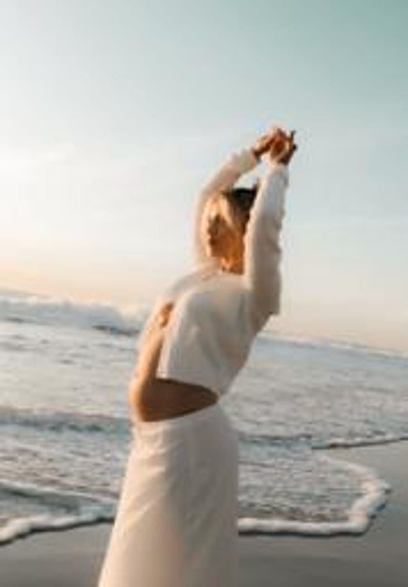 K&ouml;nnte beinhalten: Eine Frau in einem wei&szlig;en, kurz geschnittenen Oberteil mit langen &Auml;rmeln und einem passenden Rock steht an einem Strand. Die Meereswellen sind im Hintergrund unter einem hellblauen Himmel zu sehen. Die Frau hat die Arme &uuml;ber dem Kopf erhoben.