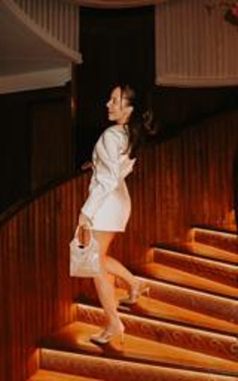 Puede incluir: Una mujer con un vestido blazer blanco y un bolso de mano blanco con una cadena plateada subiendo una escalera de madera. El vestido tiene mangas largas y un corte entallado. La escena est&aacute; iluminada con una luz c&aacute;lida. Lleva tacones altos.