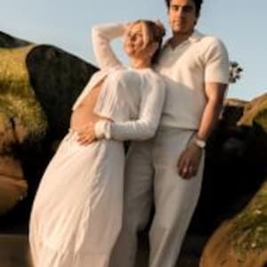 K&ouml;nnte beinhalten: Ein Paar posiert im Freien. Die Frau tr&auml;gt ein langes wei&szlig;es Kleid, der Mann ein cremefarbenes Polohemd und eine Hose. Sie stehen in der N&auml;he gro&szlig;er Felsen, wobei weiches, nat&uuml;rliches Licht die Szene erhellt.