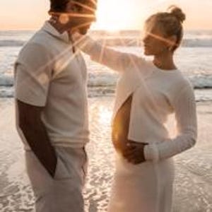 K&ouml;nnte beinhalten: Ein Paar steht am Strand bei Sonnenuntergang. Die Frau, in einem wei&szlig;en Kleid, ber&uuml;hrt sanft das Gesicht des Mannes. Der Mann tr&auml;gt ein wei&szlig;es Polohemd und eine Hose. Die Sonne scheint hinter ihnen und erzeugt einen warmen, goldenen Schein.