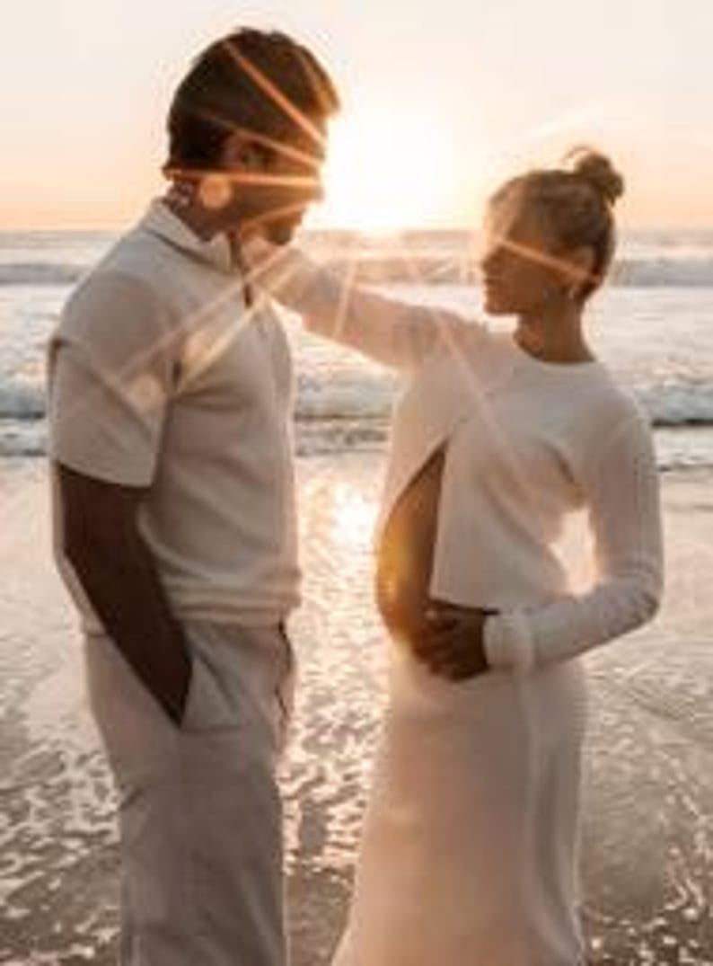 K&ouml;nnte beinhalten: Ein Paar steht am Strand bei Sonnenuntergang. Die Frau, in einem wei&szlig;en Kleid, ber&uuml;hrt sanft das Gesicht des Mannes. Der Mann tr&auml;gt ein wei&szlig;es Polohemd und eine Hose. Die Sonne scheint hinter ihnen und erzeugt einen warmen, goldenen Schein.