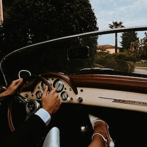 Puede incluir: Interior de un Porsche descapotable cl&aacute;sico. El salpicadero es crema con detalles de madera marr&oacute;n y medidores. Las manos est&aacute;n en el volante de madera y se ve un pie con un zapato blanco. El coche circula por una carretera arbolada.