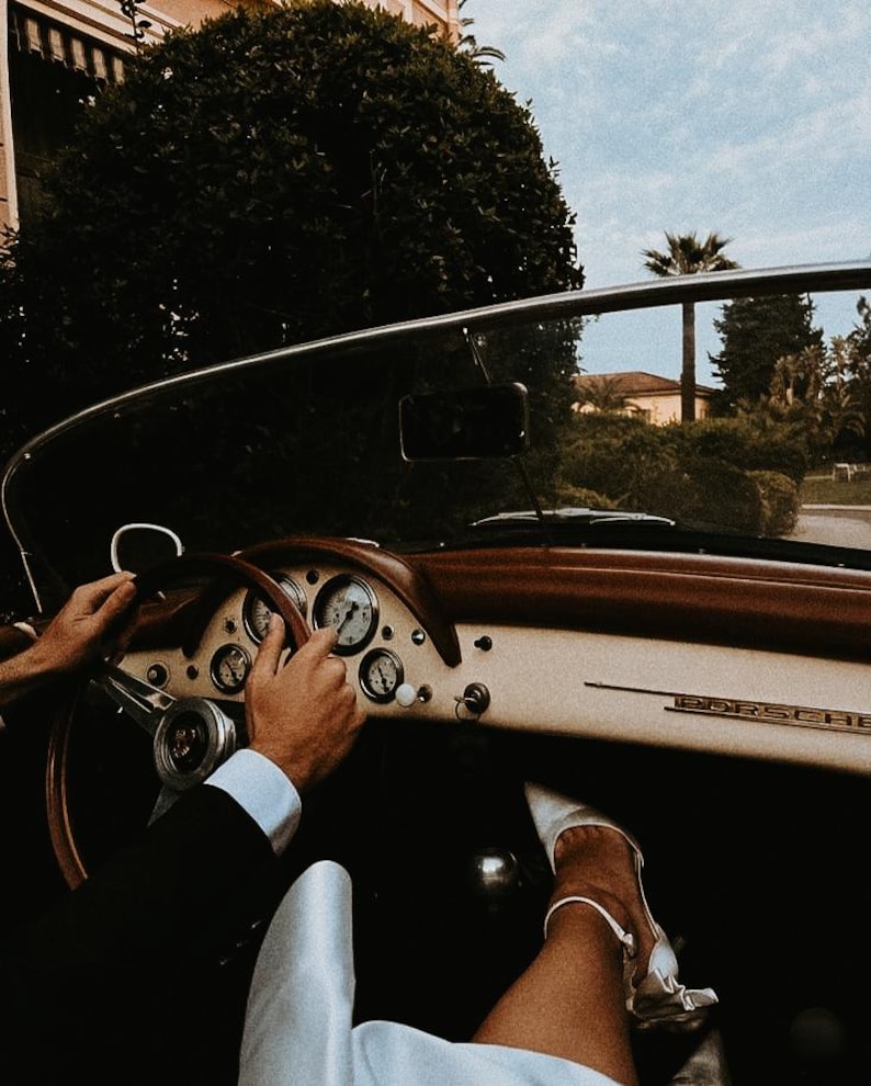 Puede incluir: Interior de un Porsche descapotable cl&aacute;sico. El salpicadero es crema con detalles de madera marr&oacute;n y medidores. Las manos est&aacute;n en el volante de madera y se ve un pie con un zapato blanco. El coche circula por una carretera arbolada.