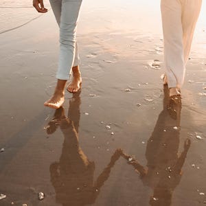 Pode incluir: Duas pessoas caminhando descal&ccedil;as em uma praia molhada ao p&ocirc;r do sol. Uma pessoa usa cal&ccedil;as azul claro, a outra cal&ccedil;as brancas. Suas sombras s&atilde;o projetadas na areia molhada, criando uma imagem espelhada.