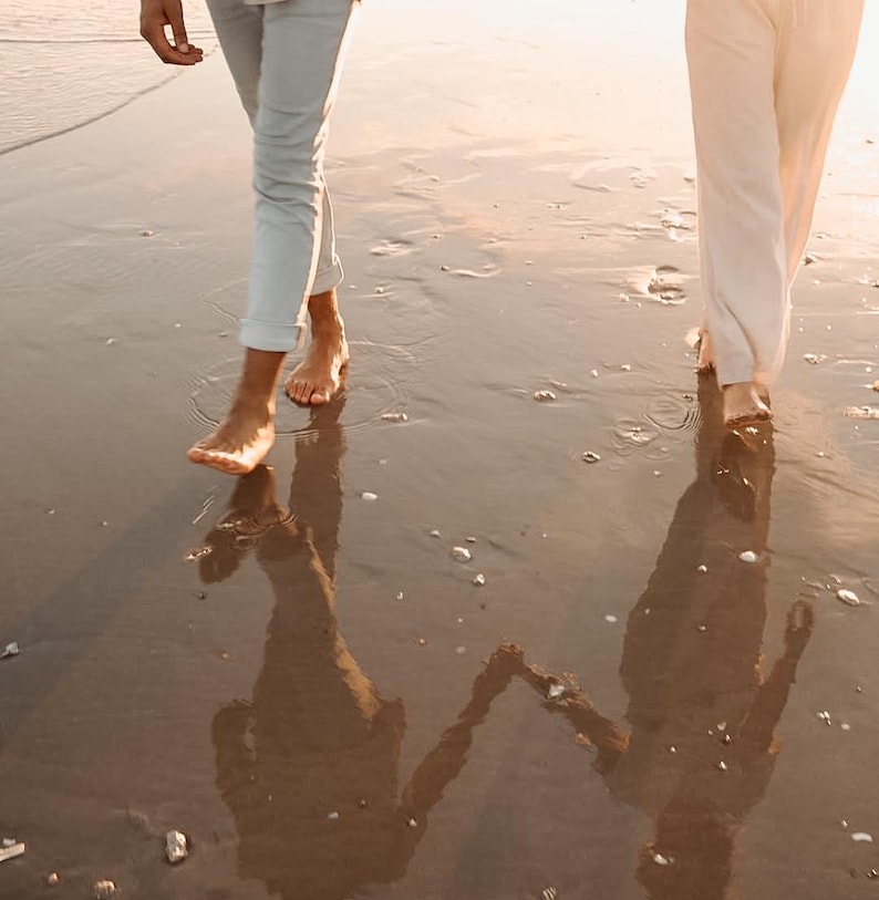 Pode incluir: Duas pessoas caminhando descal&ccedil;as em uma praia molhada ao p&ocirc;r do sol. Uma pessoa usa cal&ccedil;as azul claro, a outra cal&ccedil;as brancas. Suas sombras s&atilde;o projetadas na areia molhada, criando uma imagem espelhada.