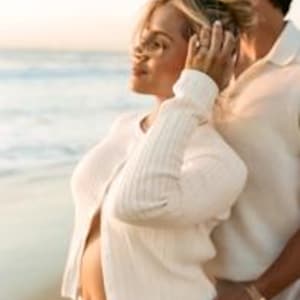 K&ouml;nnte beinhalten: Eine schwangere Person tr&auml;gt eine wei&szlig;e Strickjacke und eine wei&szlig;e Hose an einem Strand, mit dem Meer im Hintergrund. Eine andere Person steht hinter ihr, mit dem Arm um die Taille der schwangeren Person.