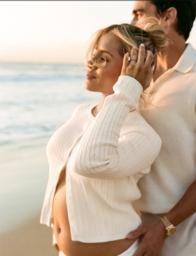 K&ouml;nnte beinhalten: Eine schwangere Person tr&auml;gt eine wei&szlig;e Strickjacke und eine wei&szlig;e Hose an einem Strand, mit dem Meer im Hintergrund. Eine andere Person steht hinter ihr, mit dem Arm um die Taille der schwangeren Person.