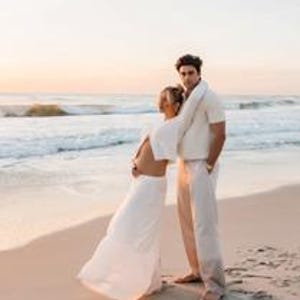 K&ouml;nnte beinhalten: Ein Paar steht am Strand bei Sonnenuntergang. Die Frau tr&auml;gt ein wei&szlig;es Kleid mit flie&szlig;endem Rock und offenem R&uuml;cken. Der Mann tr&auml;gt ein cremefarbenes kurz&auml;rmeliges Hemd und eine passende Hose. Sanfte Wellen umsp&uuml;len das Ufer.