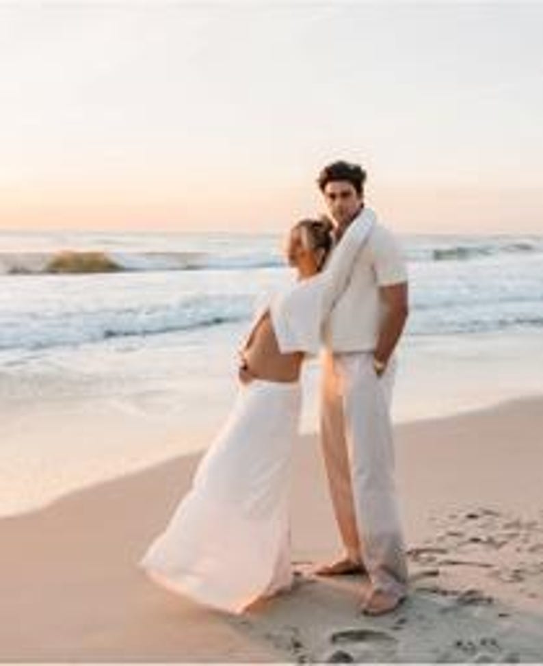 K&ouml;nnte beinhalten: Ein Paar steht am Strand bei Sonnenuntergang. Die Frau tr&auml;gt ein wei&szlig;es Kleid mit flie&szlig;endem Rock und offenem R&uuml;cken. Der Mann tr&auml;gt ein cremefarbenes kurz&auml;rmeliges Hemd und eine passende Hose. Sanfte Wellen umsp&uuml;len das Ufer.