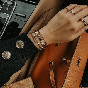 Puede incluir: Un bolso de mano de cuero marr&oacute;n claro con herrajes dorados se sostiene en un coche. La persona lleva pulseras y anillos de oro. Se ve la manga de un jersey negro con botones dorados.